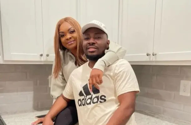 A woman and man lean on a kitchen counter, smiling at the camera, with white cabinets in the background. The man wears an Adidas shirt and a cap; the woman rests her arm on his shoulder.