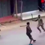 Three men in a street brawl: one in dark clothing faces two others, each wielding a wooden stick, on a sidewalked urban road.