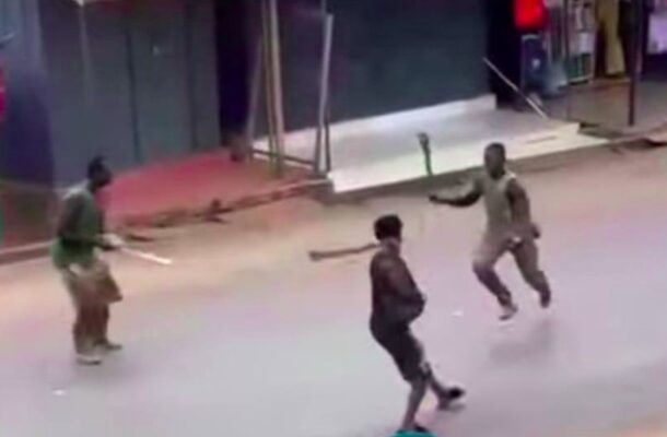 Three men in a street brawl: one in dark clothing faces two others, each wielding a wooden stick, on a sidewalked urban road.