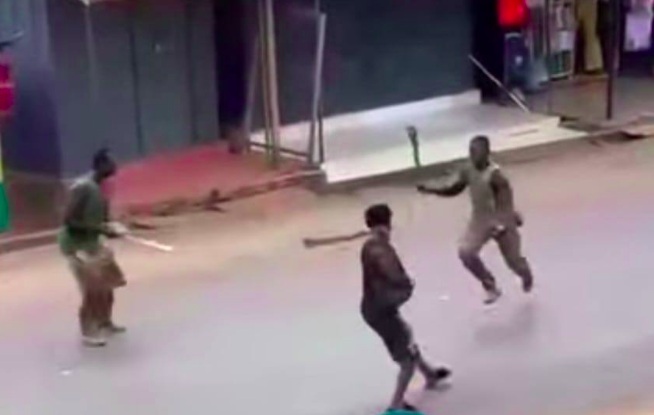 Three men in a street brawl: one in dark clothing faces two others, each wielding a wooden stick, on a sidewalked urban road.