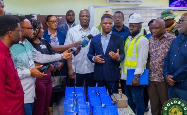 Group of officials and reporters gather for a press briefing as a man in a navy suit speaks, surrounded by workers and blue bags on the table.