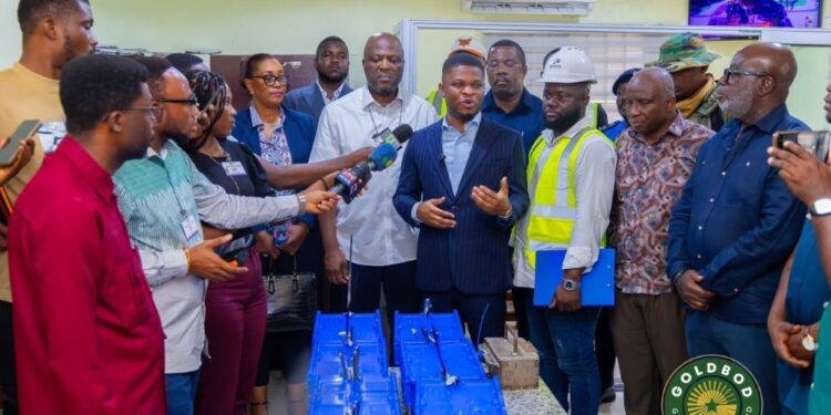 Group of officials and reporters gather for a press briefing as a man in a navy suit speaks, surrounded by workers and blue bags on the table.