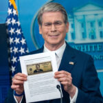 Man in a navy suit holds a printed document showing a U.S. dollar bill at a White House briefing backdrop.