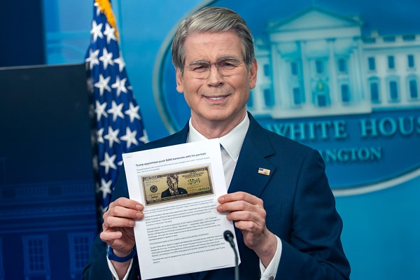 Man in a navy suit holds a printed document showing a U.S. dollar bill at a White House briefing backdrop.
