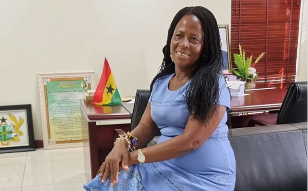 doryoe | Ghanaguardian.com Smiling woman in a light blue dress seated on a gray sofa in an office, with a small Ghana flag and framed certificates in the background.