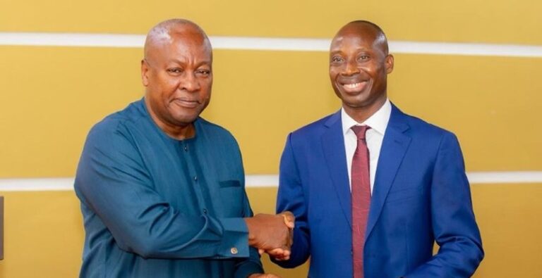Two men shake hands and smile during a formal meeting; one wears a blue traditional outfit, the other a blue suit and red tie, against a yellow wall.
