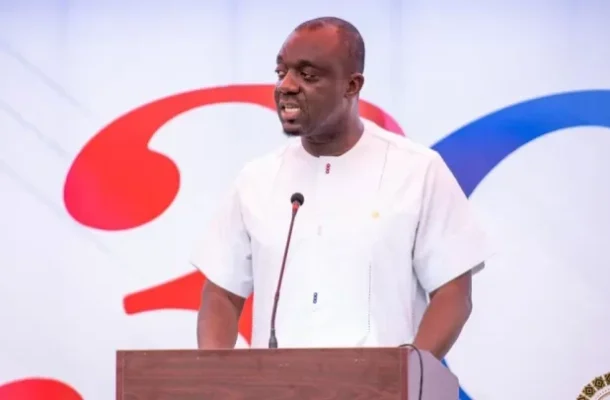 Man in a white shirt speaks at a podium with a microphone on a stage backdrop featuring red and blue shapes.
