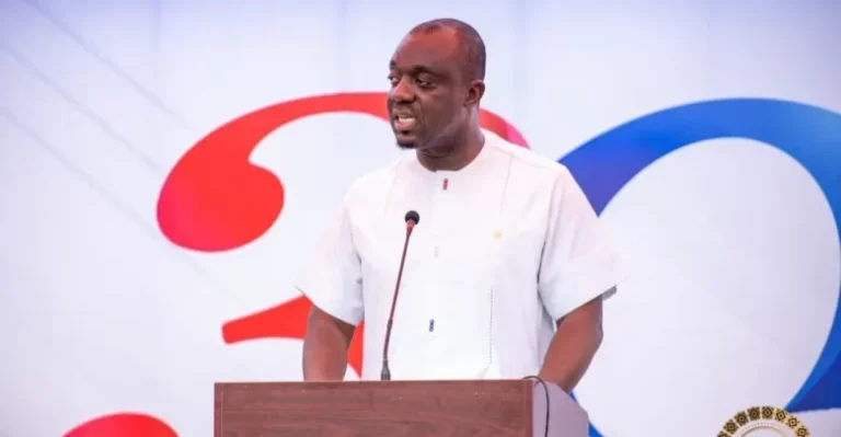 Man in a white shirt speaks at a podium with a microphone on a stage backdrop featuring red and blue shapes.