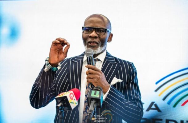 Male speaker in a pinstripe suit and glasses, holding a microphone and gesturing, at a press event with multiple microphones and an Africa Prosperity Network backdrop
