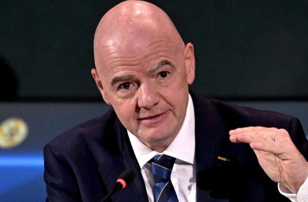 Bald man in a dark suit speaking into a microphone while gesturing with his hand at a panel.