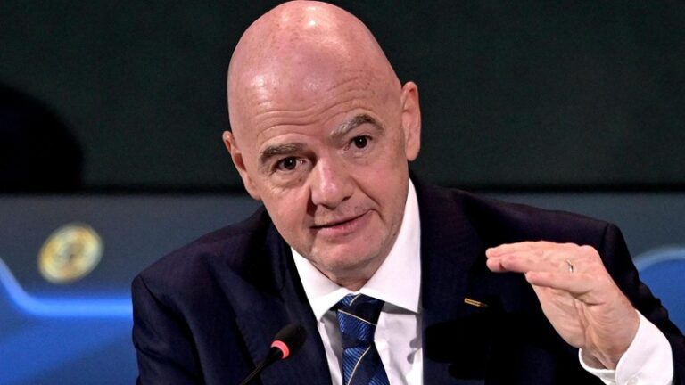 Bald man in a dark suit speaking into a microphone while gesturing with his hand at a panel.