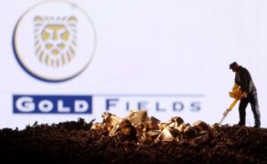 Miniature figurine miner digging in soil with gold nuggets scattered, Gold Fields logo in the background.