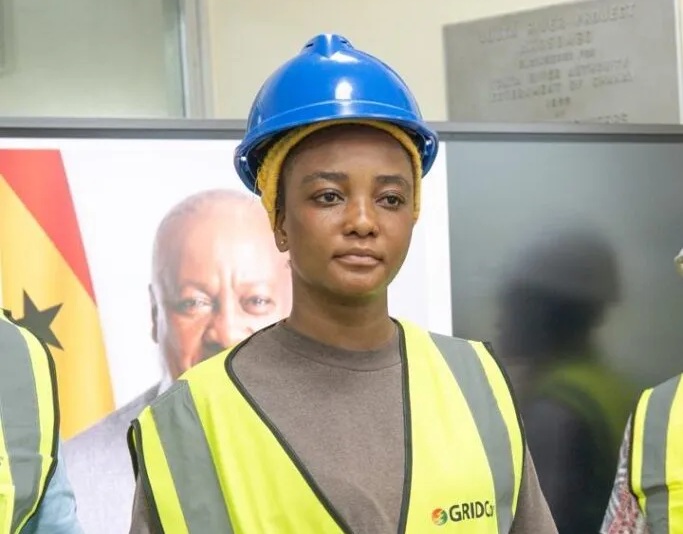 Woman construction worker wearing a blue hard hat and neon safety vest, standing indoors with a banner in the background.