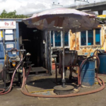 Old outdoor fueling area with tangled hoses, metal drums, and a rusted shelter under a large umbrella