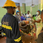 Emergency responders in PPE tend to a patient on a hospital bed with an IV drip in a treatment room.