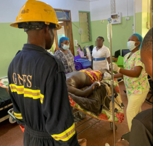 Emergency responders in PPE tend to a patient on a hospital bed with an IV drip in a treatment room.