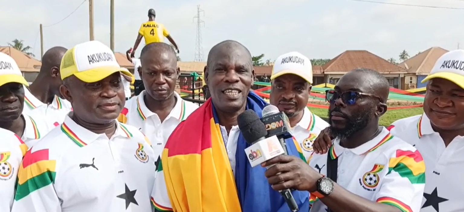 Group of Ghanaian men in white polo shirts with colorful stripes surround a man draped in flags, giving a press interview with microphones