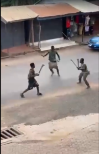 Three boys on a street, each holding a stick as if in a mock fight, near a row of shopfronts behind them.