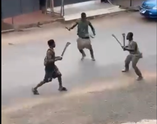 Three boys on a street, each holding a stick as if in a mock fight, near a row of shopfronts behind them.