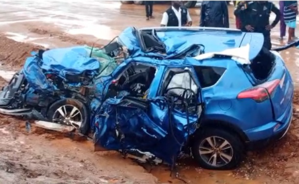 Blue SUV severely damaged in a high-impact collision on a muddy road, with the front end crushed and doors mangled; onlookers stand nearby.