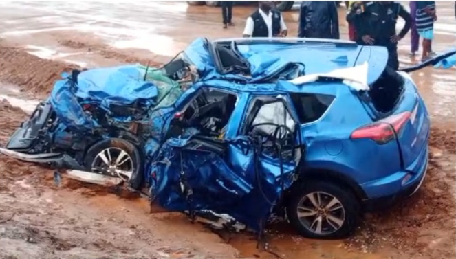 Blue SUV severely damaged in a high-impact collision on a muddy road, with the front end crushed and doors mangled; onlookers stand nearby.