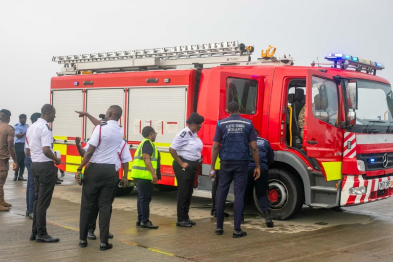Red fire engine with blue lights parked on a tarmac; several people, including uniforms, stand nearby inspecting the vehicle