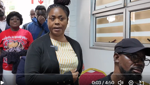 image-2721 | Ghanaguardian.com A woman in a yellow striped blouse and dark cardigan speaks to a small group inside a room, with a man wearing a cap seated to her right and others standing in the background.