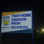 Night photo of a sign for Tanyigbe Senior High School with a crest on the left side? (Note: remove question mark)