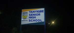 Night photo of a sign for Tanyigbe Senior High School with a crest on the left side? (Note: remove question mark)