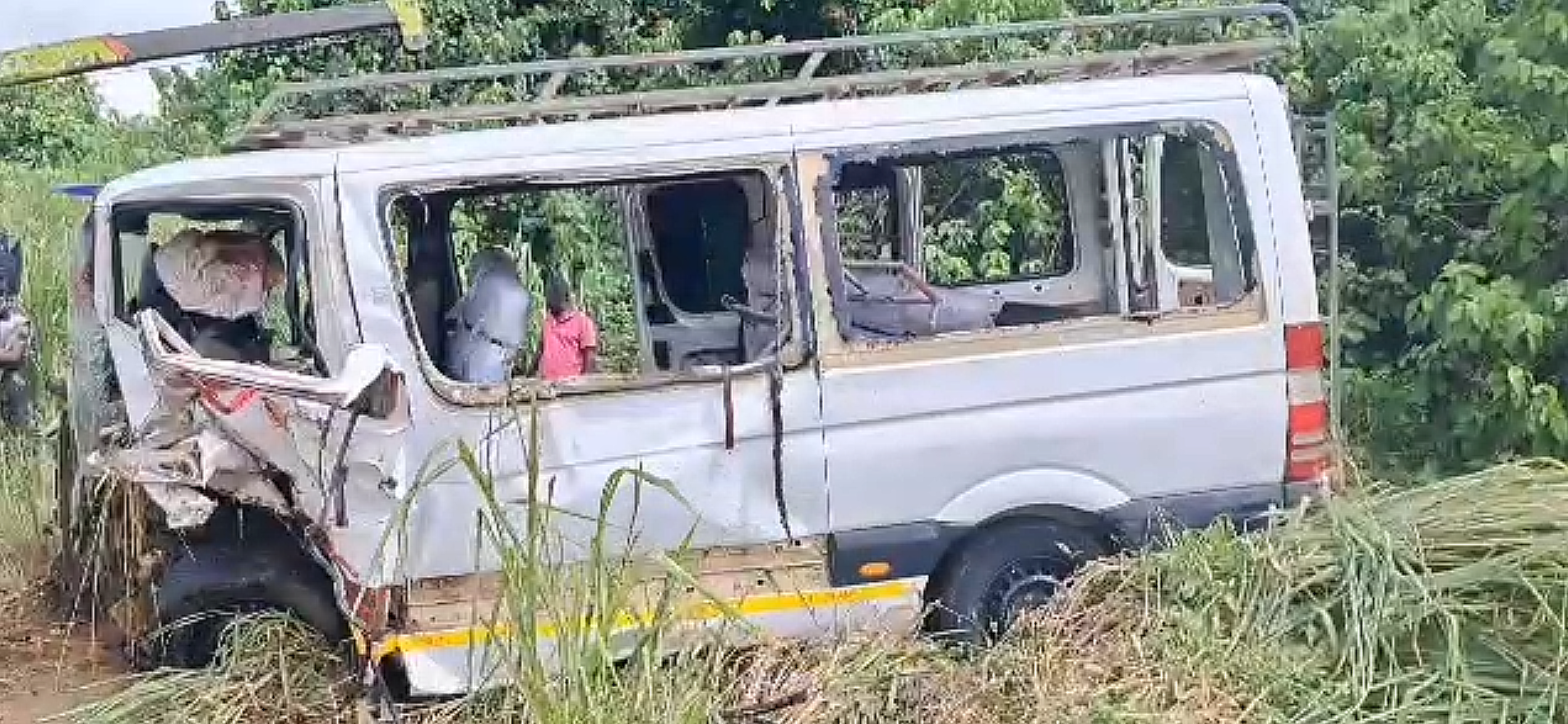 White van heavily damaged with front end crushed, drivers side on grassy ditch amid tall weeds and water nearby; appears abandoned after an accident