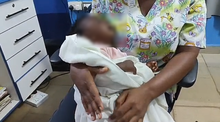 Healthcare worker in a floral top cradling a newborn wrapped in white cloth in a medical room.