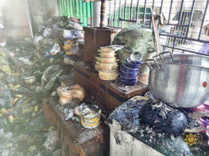 Smoke-damaged, cluttered kitchen counter with burnt containers, a large metal pot, and charred debris after a fire