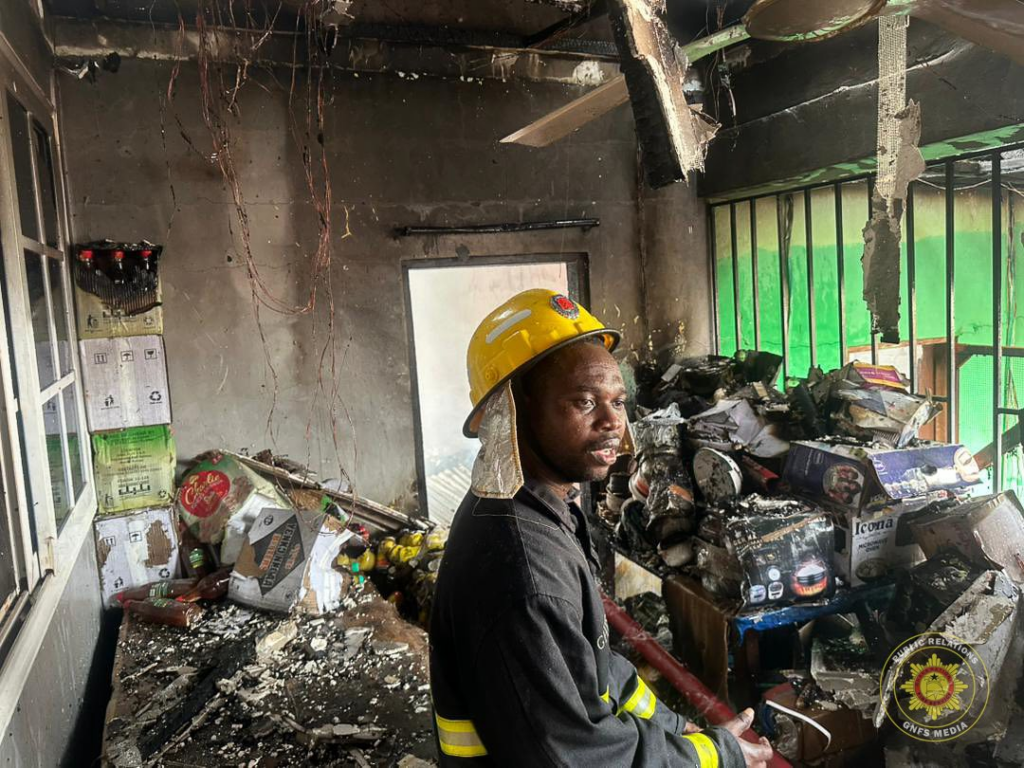 Firefighter in a yellow helmet sits among charred debris in a burned-out room.