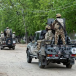 Military convoy of soldiers in camouflage gear riding in pickup trucks along a tree-lined dirt road, helmets on and armed.