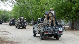 Military convoy of soldiers in camouflage gear riding in pickup trucks along a tree-lined dirt road, helmets on and armed.