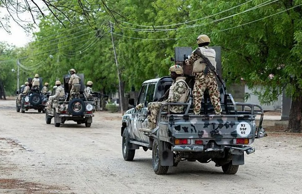 Military convoy of soldiers in camouflage gear riding in pickup trucks along a tree-lined dirt road, helmets on and armed.