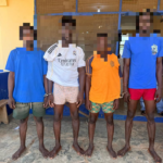 Four barefoot men stand in a line outside a yellow building with blue trim; their hands are restrained in front, faces pixelated for privacy, wearing casual shorts and t-shirts or a jersey.