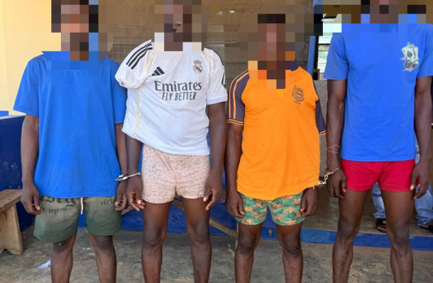 Four barefoot men stand in a line outside a yellow building with blue trim; their hands are restrained in front, faces pixelated for privacy, wearing casual shorts and t-shirts or a jersey.