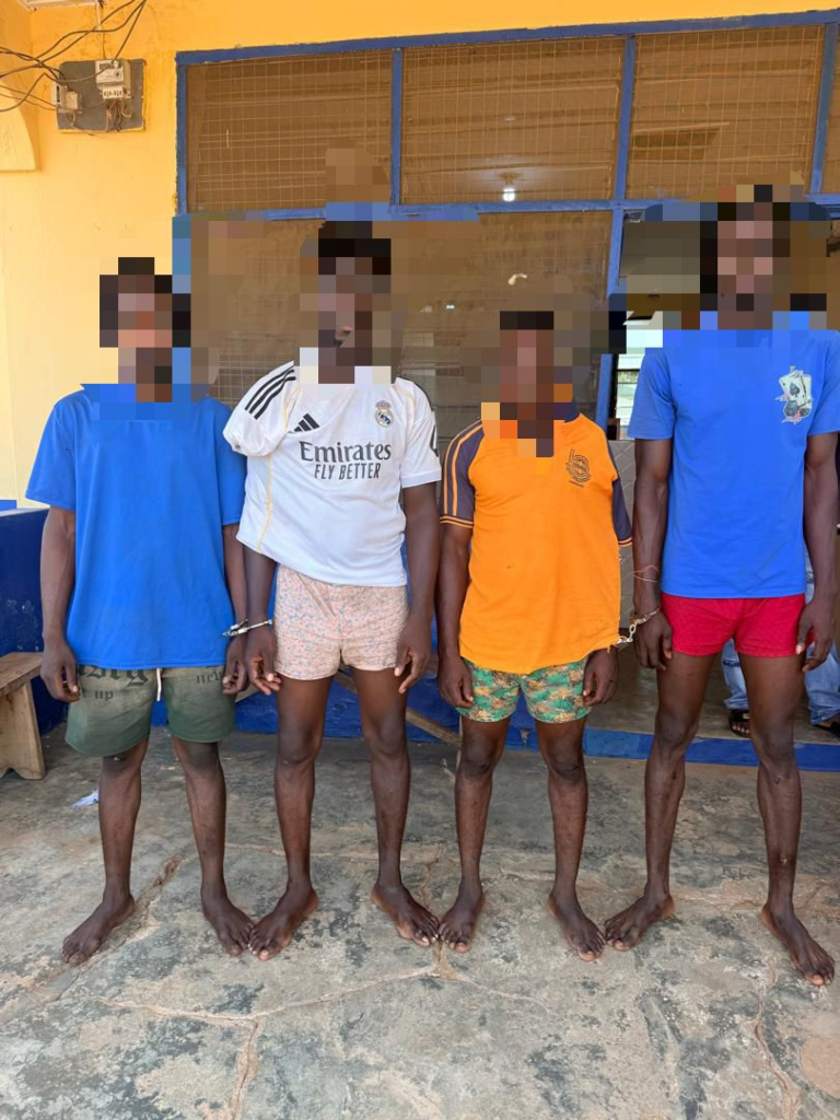 Four barefoot men stand in a line outside a yellow building with blue trim; their hands are restrained in front, faces pixelated for privacy, wearing casual shorts and t-shirts or a jersey.
