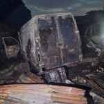 Charred van and debris in a burned-out junkyard at dusk, with a smoky sky above.