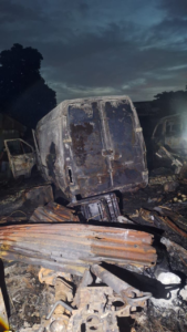 Charred van and debris in a burned-out junkyard at dusk, with a smoky sky above.