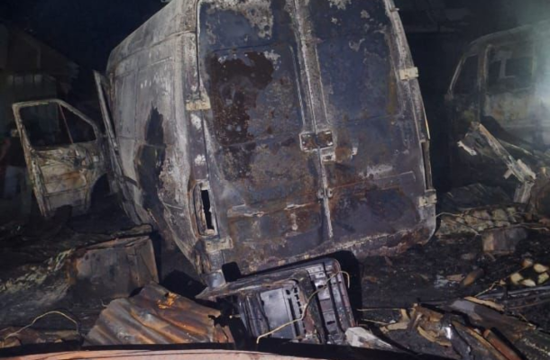Charred van and debris in a burned-out junkyard at dusk, with a smoky sky above.