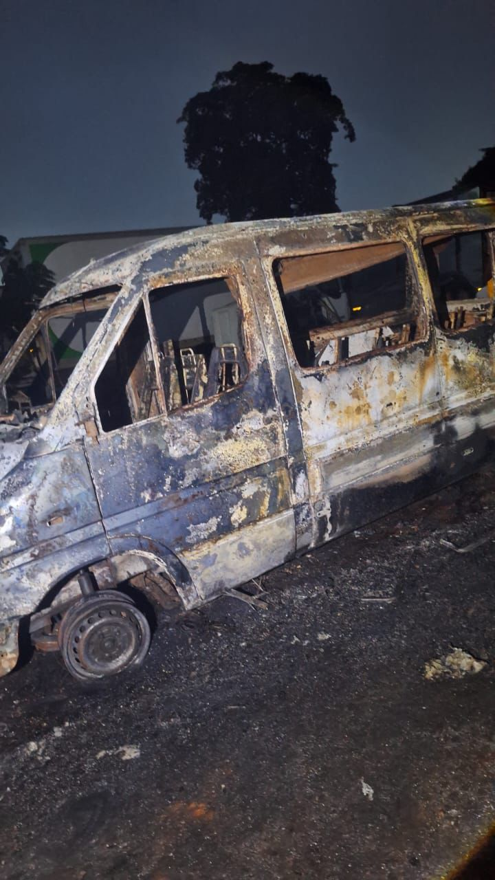 Burned-out van with extensive fire damage, shattered windows, and charred exterior at night on a scorched lot