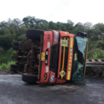 Orange bus overturned on its side on a rural road, with broken windshield and debris nearby.