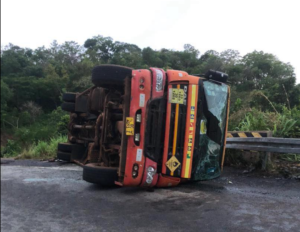 Orange bus overturned on its side on a rural road, with broken windshield and debris nearby.