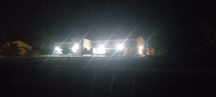 Night exterior of a single-story building with bright white lights along the façade, surrounded by darkness and trees/smaller structures nearby.