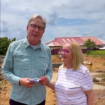 Two adults stand at a dirt construction site, the man holding papers while the woman smiles beside him, with a red-roofed building in the background.