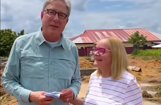 Two adults stand at a dirt construction site, the man holding papers while the woman smiles beside him, with a red-roofed building in the background.