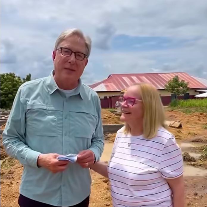 Two adults stand at a dirt construction site, the man holding papers while the woman smiles beside him, with a red-roofed building in the background.