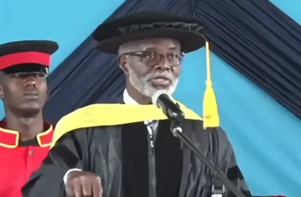 Elderly man in a black graduation gown and cap speaks at a podium, yellow stole draped over shoulders, microphone in front.